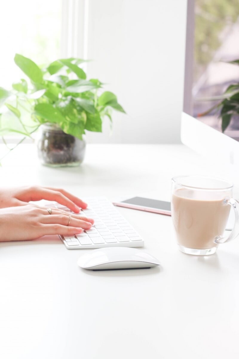 A pretty home office with bright white colors. A close up of someones hands working on a computer at their desk.