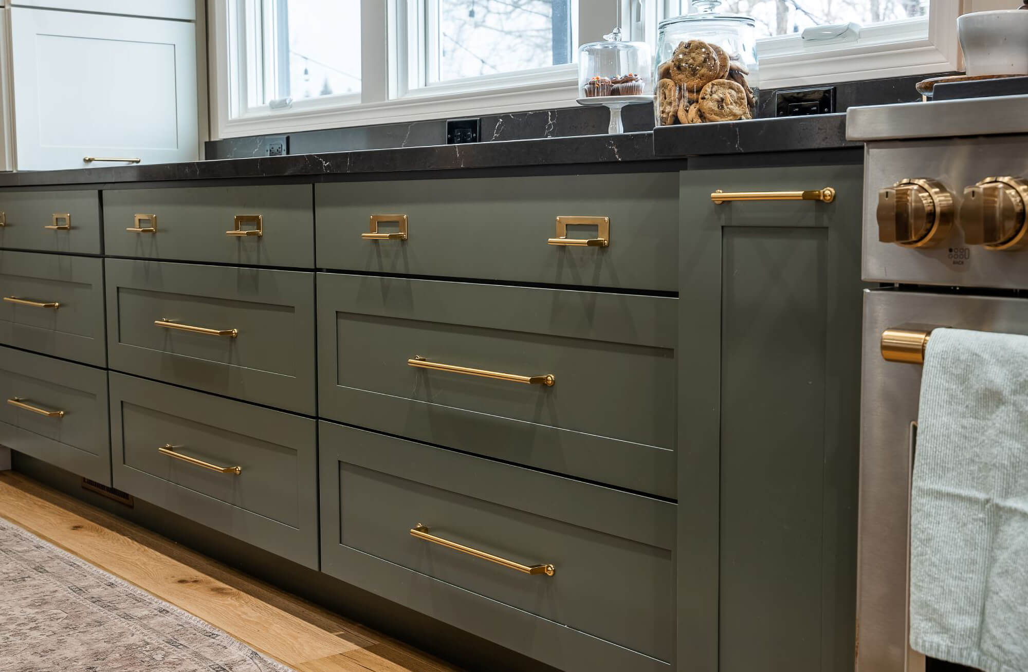 A close up of the beautiful dark green painted cabinets with a shallow, skinny shaker door style with gold, brassy hardware.